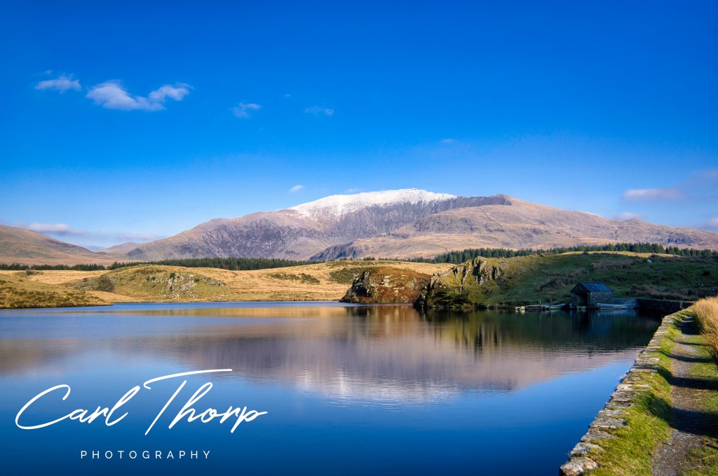 Winter view of Yr Wddfa (Snowdon) capped with snow and blue skys reflecting in a lake.